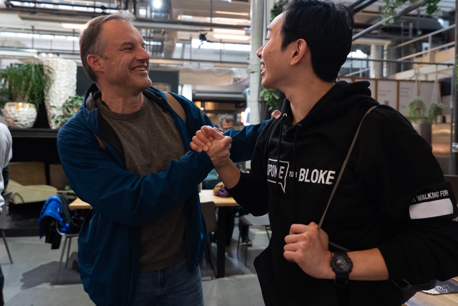 Two men sharing a warm handshake and a laugh, one wearing a Spoke to a Bloke hoodie