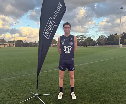 Dane Plakalovic standing beside a Spoke to a Bloke flag on a football field
